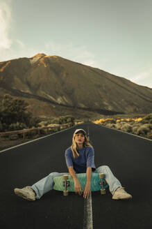 Blonde girl with a cap sitting in the middle of the road posing with a Skateboard with the Teide in the background. Tenerife, Canary Islands, Spain. - ACPF01580