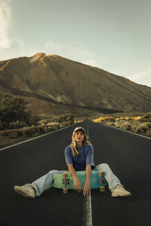 Carefree woman sitting with skateboard on mountain road in front of Mount Teide - ACPF01580