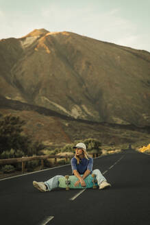 Blonde girl with a cap sitting in the middle of the road posing with a Skateboard with the Teide in the background. Tenerife, Canary Islands, Spain. - ACPF01581