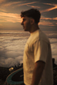 Boy holding a skateboard with a nice sea of clouds in the background during sunset. Tenerife, Canary Islands, Spain., blurred - ACPF01582