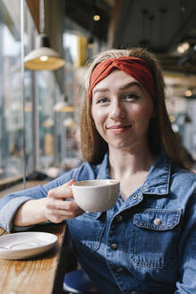 Lächelnde junge Frau sitzt mit einer Kaffeetasse in einem Café - ASGF04983