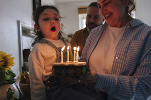 Glücklicher Vater und Mutter mit Tochter blasen Kerzen auf Geburtstagskuchen zu Hause - ASGF05024