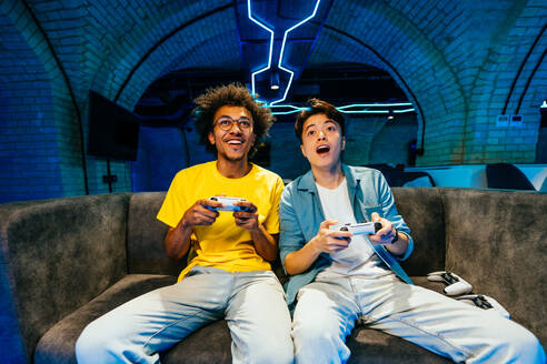 Multiethnic group of young friends playing videogames at home sitting on the sofa - Videogamers having fun playing e-sports at computer videogame console - DMDF11698