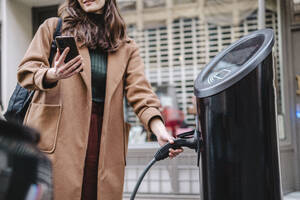Geschäftsfrau mit Smartphone, die einen Stecker an einer Ladestation für Fahrzeuge hält - AMWF02179