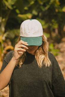 Blonde girl with a cap in front of some banana trees looking down. Tenerife, Canary Islands, Spain - ACPF01600