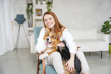 Happy woman embracing dog sitting in chair at home - NLAF00427