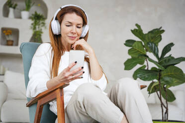 Smiling woman listening music through headphones and using smart phone at home - NLAF00429