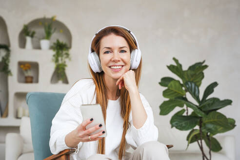 Happy woman holding smart phone listening music through headphones at home - NLAF00432