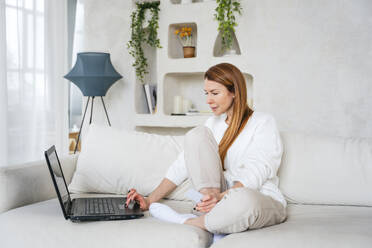 Woman holding credit card shopping online through laptop sitting on sofa at home - NLAF00437