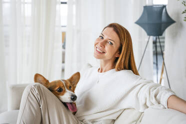 Happy woman sitting with dog in living room at home - NLAF00439