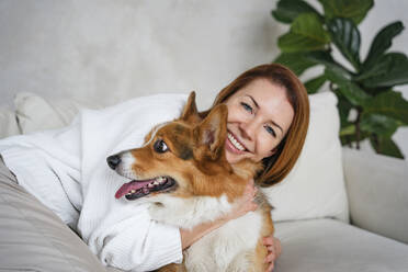Happy woman embracing dog on sofa at home - NLAF00441