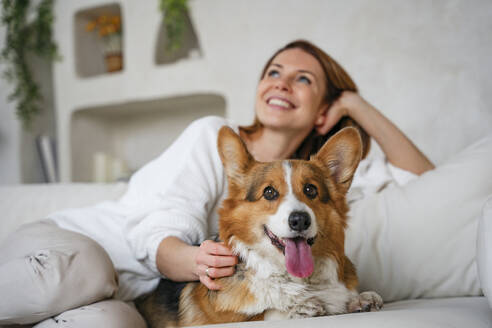Happy woman sitting with dog on couch at home - NLAF00442