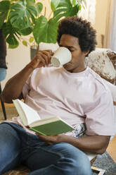 young man sitting in modern living room, drinking coffee and reading a book - FLMZF00044