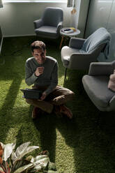 Glücklicher Geschäftsmann sitzt mit Tablet-PC auf grünem Teppich - JOSEF24105