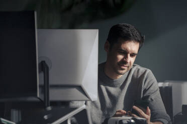 Businessman using mobile phone near desktop PC in office - JOSEF24123