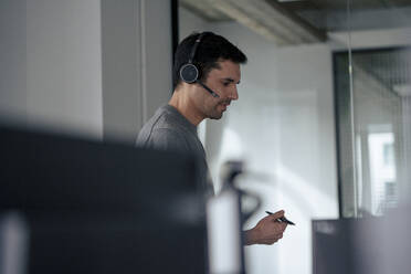 Mature businessman wearing headset and holding pen in office - JOSEF24172