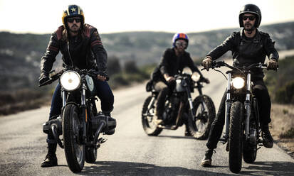 Drei Männer mit offenen Sturzhelmen und Schutzbrillen sitzen auf Cafe-Racer-Motorrädern auf einer Landstraße. - MINF16705