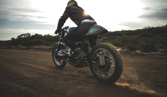 Rückansicht eines Mannes mit Sturzhelm auf einem Cafe-Racer-Motorrad auf einem staubigen Feldweg. - MINF16707