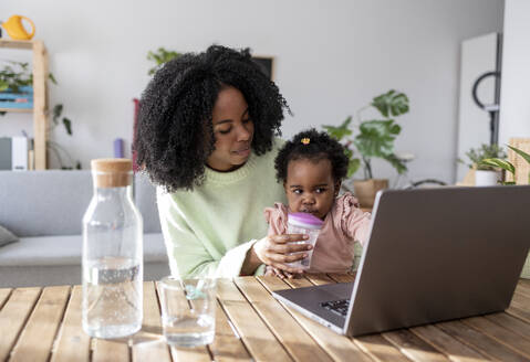 Alleinerziehende berufstätige Mutter, die ihrer Tochter beim Trinken von Wasser aus einer Babyschale hilft, während sie mit einem Laptop am Tisch sitzt - JCCMF11723