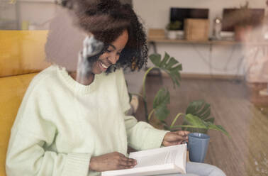 Lächelnde junge Frau mit Kaffeetasse auf einem Stuhl sitzend und ein Buch lesend, gesehen durch ein Fensterglas - JCCMF11746