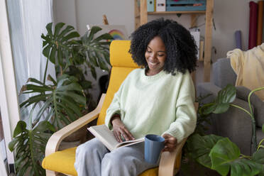 Lächelnde junge Frau sitzt auf einem Stuhl mit Kaffeetasse und liest ein Buch zu Hause - JCCMF11747