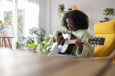 Smiling young woman with curly hair practicing guitar at home - JCCMF11761