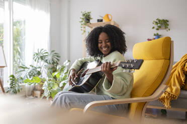 Happy young woman with curly hair practicing guitar at home - JCCMF11762