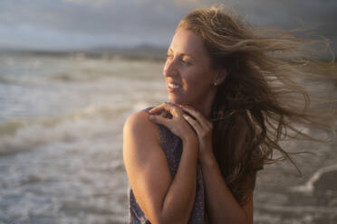 Smiling blond woman enjoying sunset at windy beach - NJAF00973