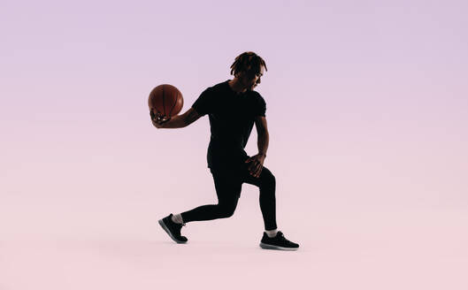 Silhouetted against a basketball court background, a young athlete with dreadlocks practices dribbling a ball with perfect form. He wears sportswear and a pink shirt. Ideal for fitness, exercise, and sports training themes. - JLPSF31655