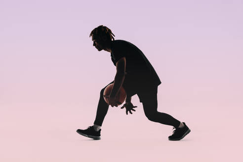Silhouetted against a pink background, a young male basketball player with dreadlocks demonstrates precision and control as he dribbles the ball. Basketball athlete training and practicing offence in a studio. - JLPSF31656