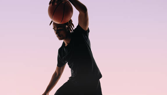 Young athlete with dreadlocks dribbles a basketball in a studio. His form is on point as he plays offense, displaying skill and athleticism. Sporting sportswear, he embodies a fit and active lifestyle, practicing diligently for the game. - JLPSF31658