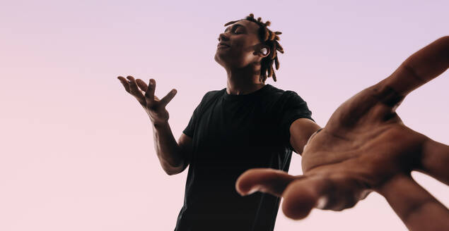 Man with dreadlocks dances energetically to music playing on his wireless earbuds. He showcases his unique dance moves in a studio, with his eyes closed and his hands in motion. - JLPSF31670