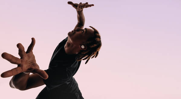 Expressive gen z male dancer with dreadlocks moves energetically in a studio, listening to music on wireless earbuds. His artistic dance moves and dynamic hand gestures bring rhythm and vibrancy to the pink silhouette. - JLPSF31675