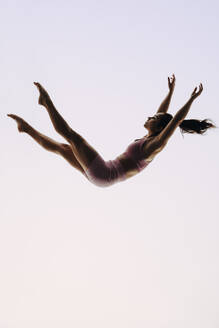 Professionelle Gymnastin in Fitnesskleidung zeigt in einem Studio ihre Beweglichkeit und Geschicklichkeit. Mit perfekter Technik vollführt sie akrobatische Kunststücke in der Luft und stellt dabei ihren flexiblen und durchtrainierten Körper zur Schau. - JLPSF31722