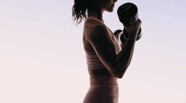 Athletic woman in sportswear does a full body workout routine in a studio. Silhouetted against a pink background, she uses dumbbells and weightlifting equipment to build muscle and improve her physique. - JLPSF31727