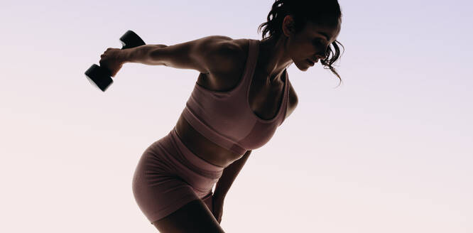 Eine sportliche Frau hebt in einem Studio Gewichte, wobei der Schwerpunkt auf Muskelaufbau und Krafttraining liegt. Eine Sportlerin trainiert mit Hanteln für ein Ganzkörpertraining und setzt sich für einen gesunden Lebensstil ein. - JLPSF31730