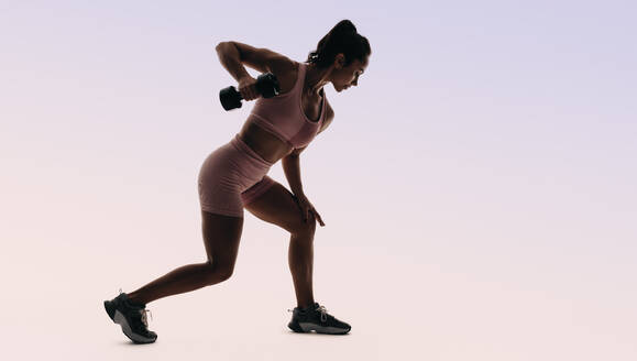 Athletic woman engaging in a workout routine in a studio, determined to crush her fitness goals. Dressed in sportswear, she lifts a dumbbell to building strength and muscles. - JLPSF31734