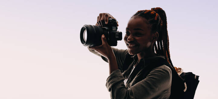 Eine Frau mit Zöpfen hält mit ihrer Digitalkamera Momente fest, während sie in einem Studio einen Rucksack trägt. Sie verkörpert die Freude an der Fotografie, schafft Erinnerungen und fängt das Wesen ihrer Motive ein. - JLPSF31739