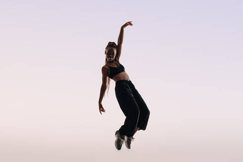 Expressive female dancer with confident posture and toned physique celebrates with dynamic moves in a studio. Fit and flexible, she embodies Gen Z's active lifestyle wearing pants and a sports bra. Full length shot. - JLPSF31801