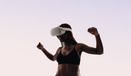 Happy, fit woman confidently celebrates winning a VR game, fully immersed in the interactive metaverse wearing a headset and goggles. Her sports bra and toned physique suggest a love for fitness, blending fun with working out. - JLPSF31806