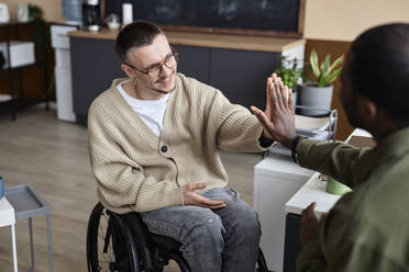 Happy businessman with disability giving high-five to colleague in office - KPEF00641