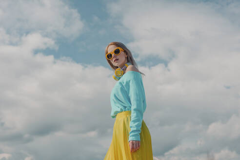 Fashionable woman wearing sunglasses and standing under cloudy sky - VSNF01863