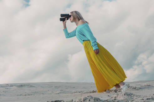 Woman wearing yellow skirt and looking through binoculars under cloudy sky - VSNF01867