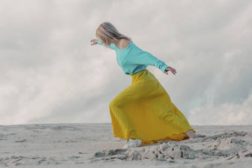 Woman wearing yellow skirt and walking on sand dune under cloudy sky - VSNF01868
