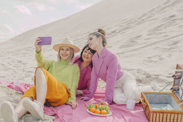 Stylish women sitting on tablecloth making selfie on picnic on sea coast, Poland - VSNF01879