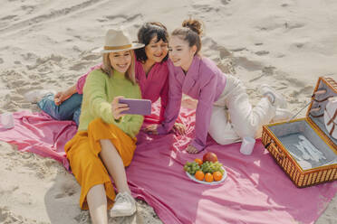 Stylish women sitting on tablecloth making selfie on picnic on sea coast, Poland - VSNF01880