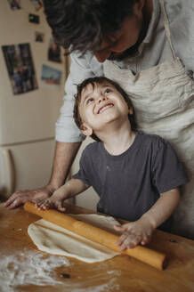 Happy son and father rolling dough in kitchen at home - ANAF02854