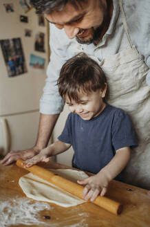 Laughing father and son rolling dough in kitchen at home - ANAF02855