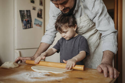 Father helping son to roll dough in kitchen at home - ANAF02856
