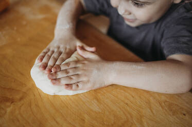 Boy pressing kneaded dough on wooden table at home - ANAF02860